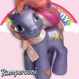 Könnte beinhalten: Lilafarbenes My Little Pony mit orangefarbenem, rosafarbenem und weißem Haar. Das Pony hat einen rosa-weißen Tupfenschal und ein Blumendesign an der Seite. Das Pony steht auf einem rosa Hintergrund mit einem Regenbogen und Wolken.
