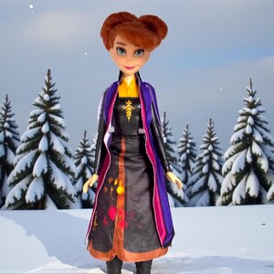 Front view of Disney Frozen II Anna Autumn Swirling Adventure doll standing in snowy forest backdrop wearing original dress, cape, and boots