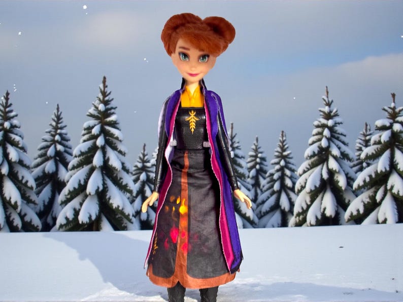 Front view of Disney Frozen II Anna Autumn Swirling Adventure doll standing in snowy forest backdrop wearing original dress, cape, and boots