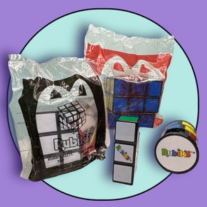 Op de afbeelding: Drie Rubik's-puzzels in verschillende maten en configuraties. De grootste puzzel is een 3x3x3-kubus met een kleurrijk ontwerp. De middelste puzzel is een 2x2x2-kubus met een wit en zwart ontwerp. De kleinste puzzel is een ronde Rubik's-bal met een wit en zwart ontwerp. De puzzels zijn verpakt in doorzichtige plastic zakken met het Rubik's-logo.