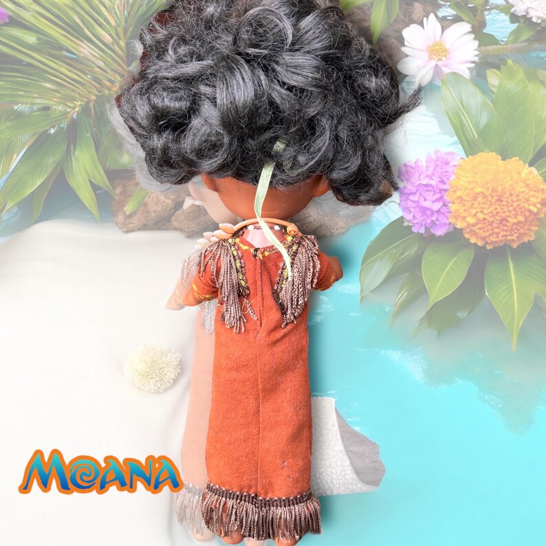 Rear view of the Moana doll, showing the back of her dress with fringe and the ribbon-tied real shell necklace. Her voluminous curls are shown from behind.