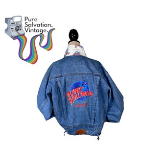 Op de afbeelding: Een blauwe denim jas met een Planet Hollywood logo-patch op de rug. De jas heeft een witte kraag met een Planet Hollywood print.