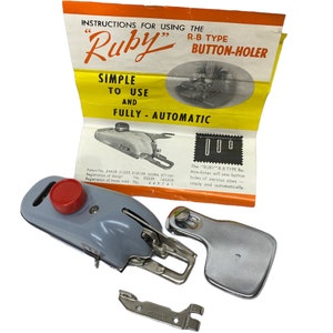 Op de afbeelding: Een grijze en zilveren knoopsgatmaker met een rode knop aan de zijkant. Het apparaat is gelabeld "Ruby" en wordt beschreven als "R-B Type Knoopsgatmaker" en "Eenvoudig te gebruiken en volledig automatisch".