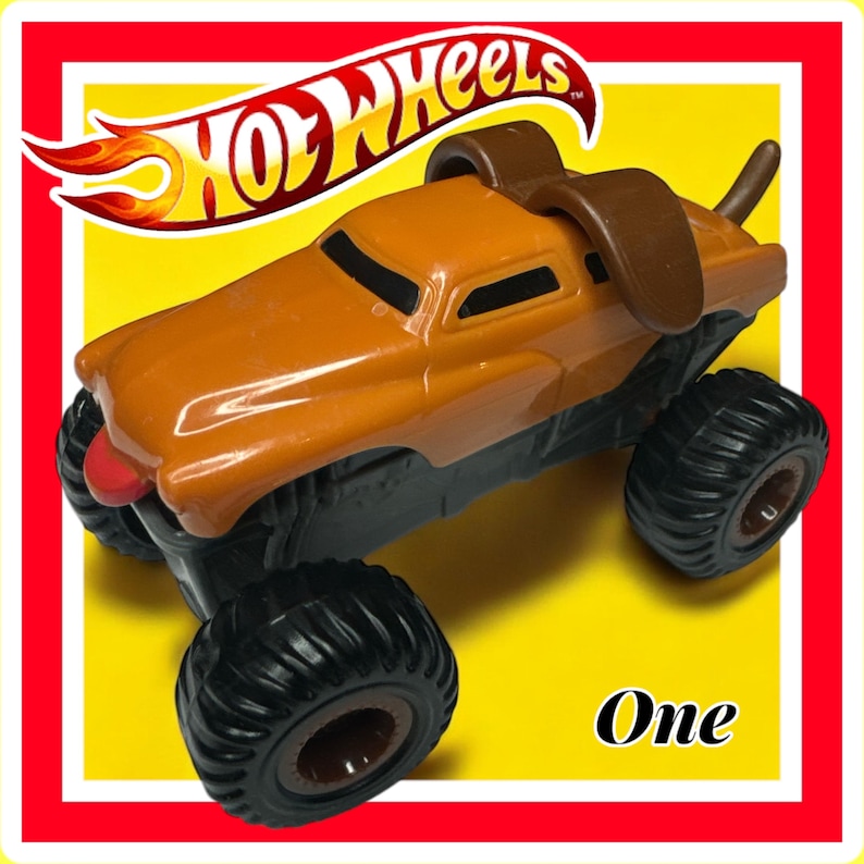 Puede incluir: Cami&oacute;n monstruo Hot Wheels naranja con una cabeza y orejas de perro marrones en la parte superior. El cami&oacute;n tiene grandes ruedas negras y una lengua roja que sobresale. El texto "One" est&aacute; impreso en el fondo amarillo.