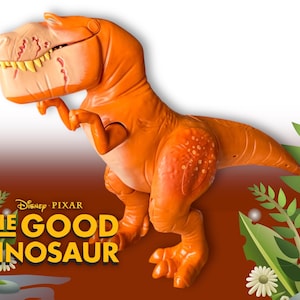 May include: An orange dinosaur toy, a character from Disney Pixar's "The Good Dinosaur." The toy has a friendly expression, visible teeth, and textured skin. The background includes the film's logo and decorative foliage.