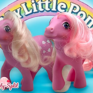 Könnte beinhalten: Zwei rosafarbene My Little Pony Spielzeuge mit langen rosa und blonden Haaren. Das Pony links hat weiße Flecken und das Pony rechts einen Regenbogenhintergrund.