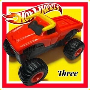 Puede incluir: Cami&oacute;n monstruo de juguete rojo con ruedas negras y una barra antivuelco amarilla. El cami&oacute;n est&aacute; sobre un fondo amarillo con las palabras "Hot Wheels" y "Three" visibles.