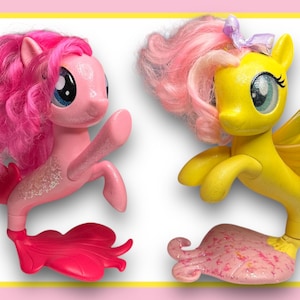 Könnte beinhalten: Zwei My Little Pony Meerjungfrauen-Figuren. Eine ist pink mit einem pinken Schwanz und die andere ist gelb mit einem pinken Schwanz.
