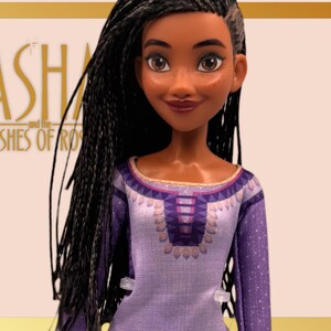 May include: A doll with long black braids and brown eyes, wearing a purple and white dress with a decorative neckline. The doll is set against a light background with the text "ASHA and the WISHES OF ROSAS".