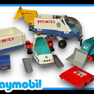May include: A set of Playmobil toys, including a white and blue space shuttle, a blue and white truck with a yellow crane arm, a blue and green construction vehicle, and a red and white storage container. The truck has the text "RYT 107 K3" on the side, and the container has the text "T 107 K3" on the side.