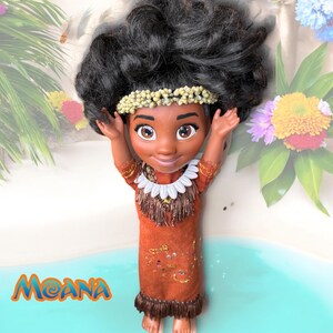 Moana doll with arms raised, smiling directly at the viewer. Her handmade island-style outfit and seashell headband are framed by a pastel beach and tropical flowers.