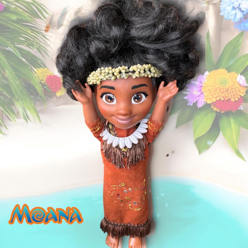 Moana doll with arms raised, smiling directly at the viewer. Her handmade island-style outfit and seashell headband are framed by a pastel beach and tropical flowers.