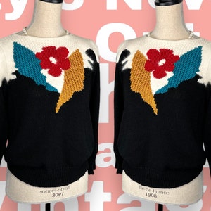 Puede incluir: Suéter de punto negro con un diseño de flor blanca, amarilla, azul y roja. El suéter tiene cuello redondo y mangas largas.