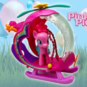 May include: Pinkie Pie, a pink pony with a pink flower in her hair, is riding in a pink helicopter with a clear bubble top. The helicopter has a rainbow design and pink hearts. The words "Pinkie Pie" are written in pink letters on a white background.