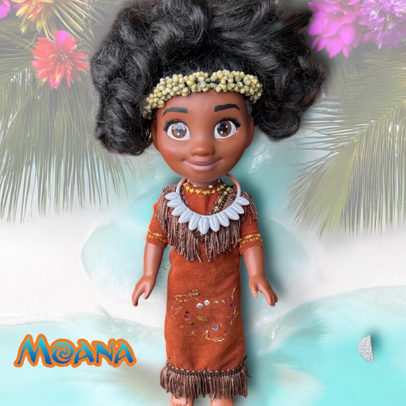 Full front view of the 14” Moana doll standing on a beach. She wears a detailed custom brown dress with beading and a white shell necklace, framed by palm leaves and flowers.