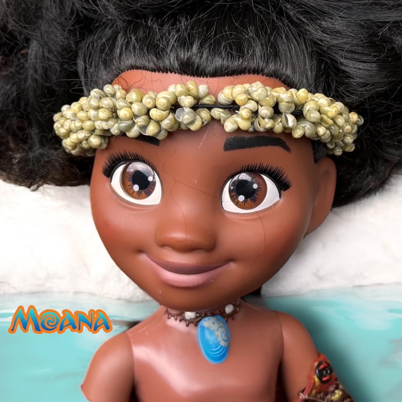 Close-up of Moana doll’s head and shoulders wearing a real ocean snail shell headband and a painted blue shell necklace. She has a warm expression and black curly hair styled up.
