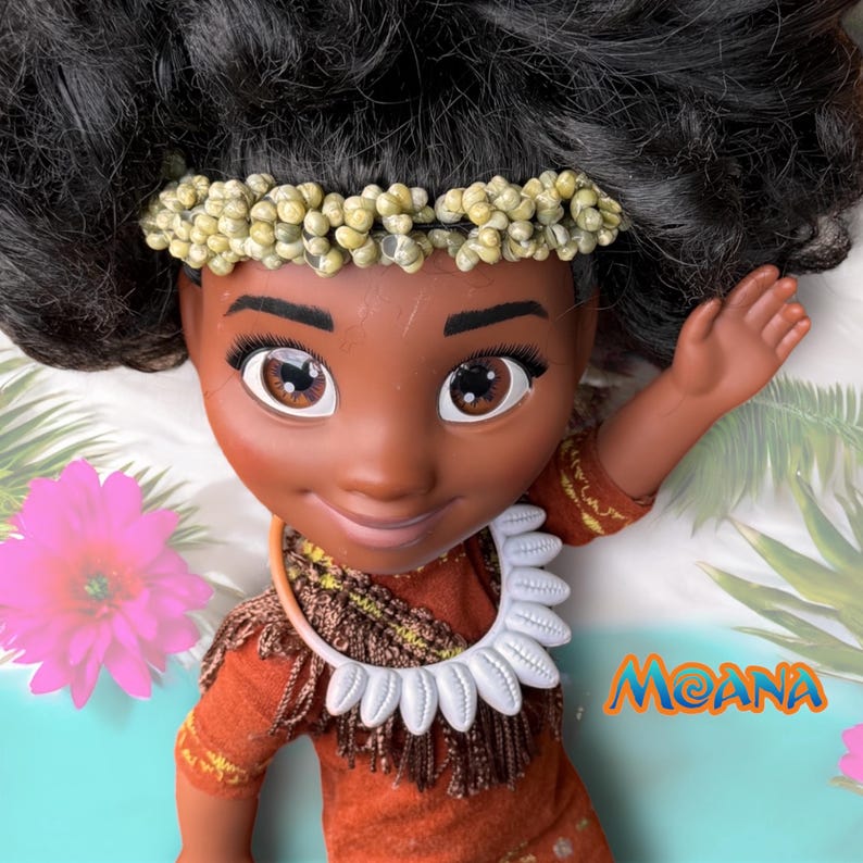Overhead view of the Moana doll smiling up, one arm raised. Her real shell headband and custom dress with fringe details are shown clearly.