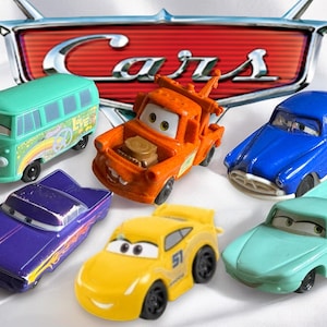 Op de afbeelding: Een verzameling van zes speelgoedauto's uit de Disney Pixar Cars-filmfranchise. De auto's zijn in verschillende kleuren, waaronder groen, oranje, blauw, paars, geel en turquoise. De auto's staan allemaal in een speelse pose, met hun wielen naar voren gericht. De auto's zijn allemaal in goede staat en lijken nieuw te zijn.