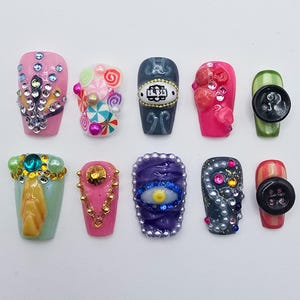 May include: A collection of ten artificial nails with various designs. The nails feature embellishments like rhinestones, candy, buttons, and intricate patterns. Colors include pink, green, blue, and gold, showcasing a range of textures and styles for nail art.