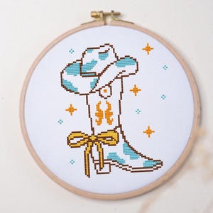 Könnte beinhalten: Eine Kreuzstichstickerei in einem runden Holzrahmen, die einen weißen Cowboystiefel mit hellblauen Kuhfleckenmustern, einem orangefarbenen flammenartigen Design und einer gelben Schleife zeigt, umgeben von kleinen orangefarbenen und blauen Sternen.