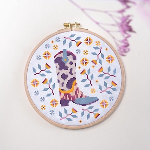 Puede incluir: Un bastidor de bordado circular de madera que contiene un patrón de punto de cruz sobre tela blanca. El diseño presenta una bota vaquera central con estampado de vaca en morado y blanco, acentuada por motivos florales en azul, amarillo y rojo.