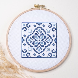 May include: A circular wooden embroidery hoop containing white fabric with a centered square cross-stitch pattern. The design features a symmetrical geometric motif in shades of dark navy blue and light powder blue, creating a tile-like appearance.