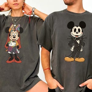 May include: Two dark gray t-shirts featuring Disney characters. One shirt displays Minnie Mouse in a colorful patchwork outfit, and the other showcases Jack Skellington from The Nightmare Before Christmas. Both shirts have short sleeves.