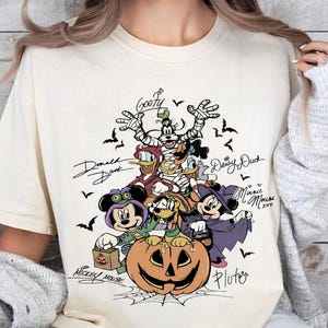 Retro-style Halloween design featuring Mickey Mouse, Minnie, Goofy, Donald, Daisy, and Pluto in costumes around a large jack-o'-lantern with bats and spiderwebs. Perfect for Disney Halloween party trips and fall-themed outfits.