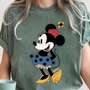 May include: A green t-shirt featuring a vintage-style image of Minnie Mouse wearing a blue polka dot skirt and yellow shoes. The shirt has the word "Disney" printed in the bottom right corner.