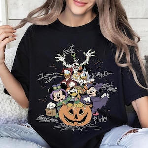 Retro-style Halloween design featuring Mickey Mouse, Minnie, Goofy, Donald, Daisy, and Pluto in costumes around a large jack-o'-lantern with bats and spiderwebs. Perfect for Disney Halloween party trips and fall-themed outfits.