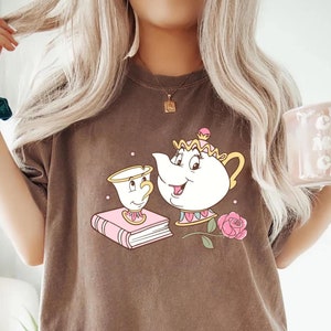 May include: A brown t-shirt with a graphic of the characters Mrs. Potts and Chip from the Disney movie Beauty and the Beast. The graphic shows Mrs. Potts holding a teacup and Chip standing next to her. The graphic is in pink, yellow, and white.