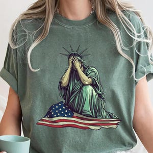 Emotional graphic of the Statue of Liberty sitting with her face in her hands wrapped in a tattered American flag symbolizing sorrow or concern for the nation perfect for political statement shirts freedom of speech tees protest art apparel or USA patriotism designs that express American values or disappointment in the state of the country
