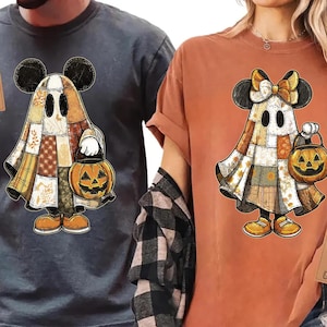 May include: Two t-shirts, one gray and one orange, each featuring a cartoon ghost character with Mickey Mouse ears and a pumpkin-shaped lantern. The ghost is dressed in a patchwork quilt costume, perfect for Halloween.