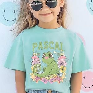 May include: A young person wearing a mint green t-shirt with the word "PASCAL" above a cartoon chameleon surrounded by flowers. The shirt is paired with blue jeans. The background features smiley face stickers.