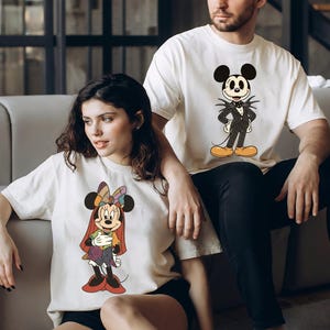 May include: Two off-white t-shirts featuring Disney characters. One shirt displays Minnie Mouse in a colorful outfit, and the other features Jack Skellington as Mickey Mouse. Both shirts are short-sleeved.