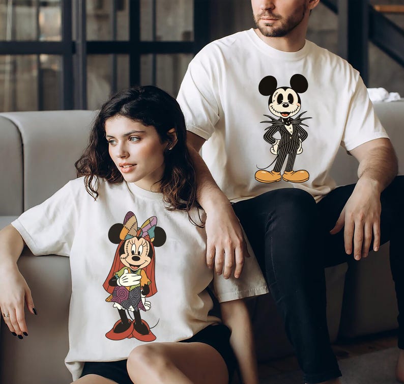 May include: Two off-white t-shirts featuring Disney characters. One shirt displays Minnie Mouse in a colorful outfit, and the other features Jack Skellington as Mickey Mouse. Both shirts are short-sleeved.