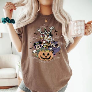 Retro-style Halloween design featuring Mickey Mouse, Minnie, Goofy, Donald, Daisy, and Pluto in costumes around a large jack-o'-lantern with bats and spiderwebs. Perfect for Disney Halloween party trips and fall-themed outfits.