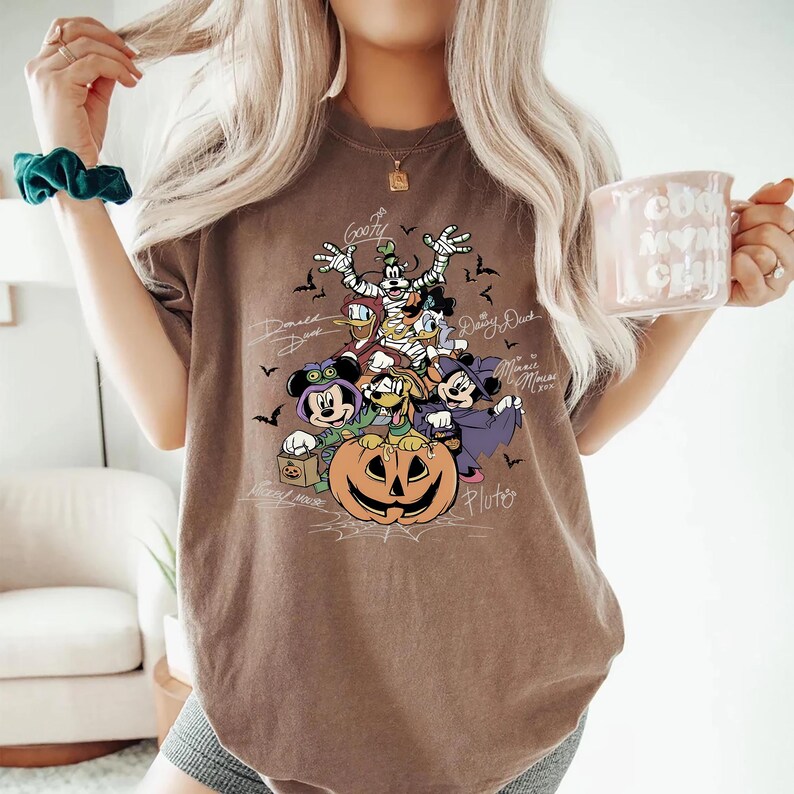Retro-style Halloween design featuring Mickey Mouse, Minnie, Goofy, Donald, Daisy, and Pluto in costumes around a large jack-o'-lantern with bats and spiderwebs. Perfect for Disney Halloween party trips and fall-themed outfits.