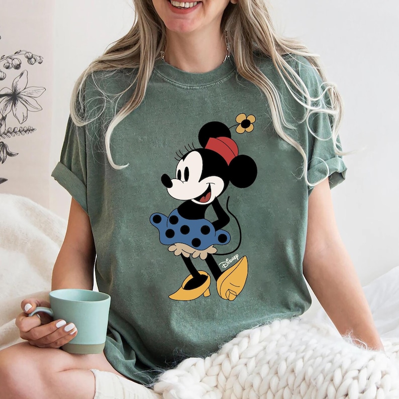 May include: A green t-shirt featuring a vintage-style image of Minnie Mouse wearing a blue polka dot skirt and yellow shoes. The shirt has the word "Disney" printed in the bottom right corner.