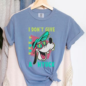 Retro 90s-style Disney shirt featuring Goofy wearing neon sunglasses and a sideways cap with bold text that says "I Don't Give A Hyuck." Colorful geometric background with a distressed vintage look. Perfect for fans of A Goofy Movie, Disney humor shirts, family vacations, or retro-themed Disney park outfits.