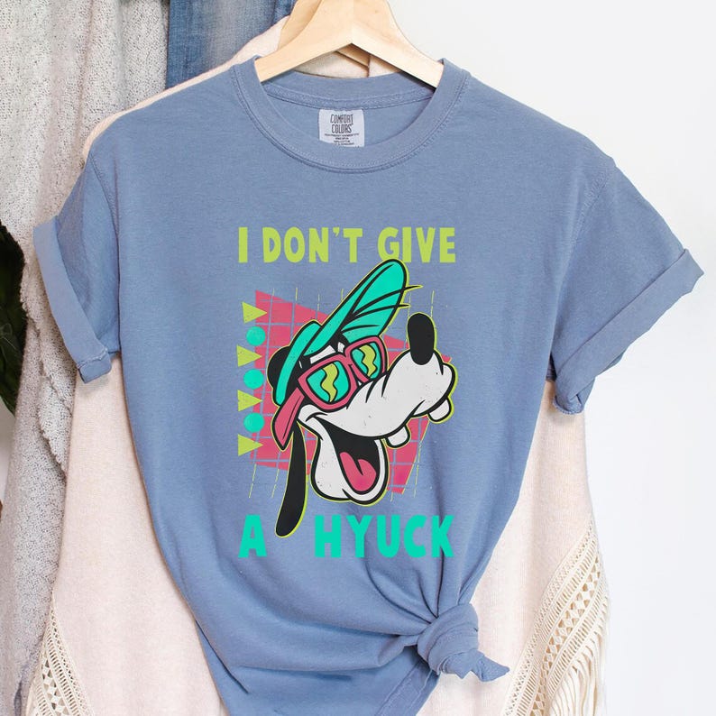 Retro 90s-style Disney shirt featuring Goofy wearing neon sunglasses and a sideways cap with bold text that says "I Don't Give A Hyuck." Colorful geometric background with a distressed vintage look. Perfect for fans of A Goofy Movie, Disney humor shirts, family vacations, or retro-themed Disney park outfits.
