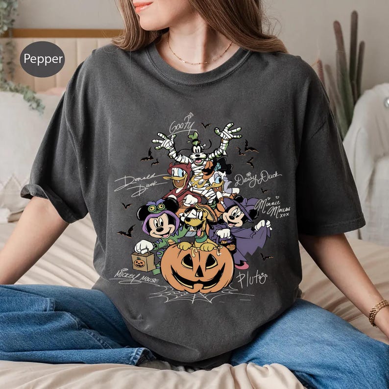 Retro-style Halloween design featuring Mickey Mouse, Minnie, Goofy, Donald, Daisy, and Pluto in costumes around a large jack-o'-lantern with bats and spiderwebs. Perfect for Disney Halloween party trips and fall-themed outfits.