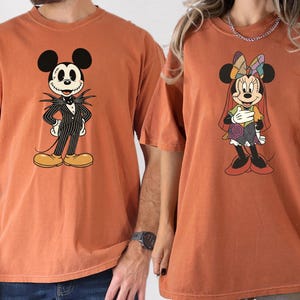 May include: Two rust-colored t-shirts featuring cartoon characters. One shirt displays Mickey Mouse dressed as Jack Skellington in a pinstripe suit. The other shirt shows Minnie Mouse in a patchwork dress. Both shirts have short sleeves.