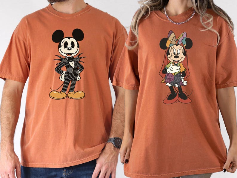 May include: Two rust-colored t-shirts featuring cartoon characters. One shirt displays Mickey Mouse dressed as Jack Skellington in a pinstripe suit. The other shirt shows Minnie Mouse in a patchwork dress. Both shirts have short sleeves.