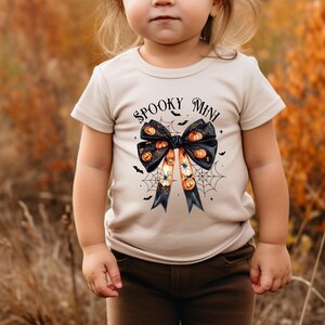 Op de afbeelding: Een peuter draagt een beige T-shirt met een zwarte strik en de tekst "Spooky Mini" erop gedrukt. De strik is versierd met oranje en zwarte pompoenen. De peuter staat in een veld met hoog bruin gras.