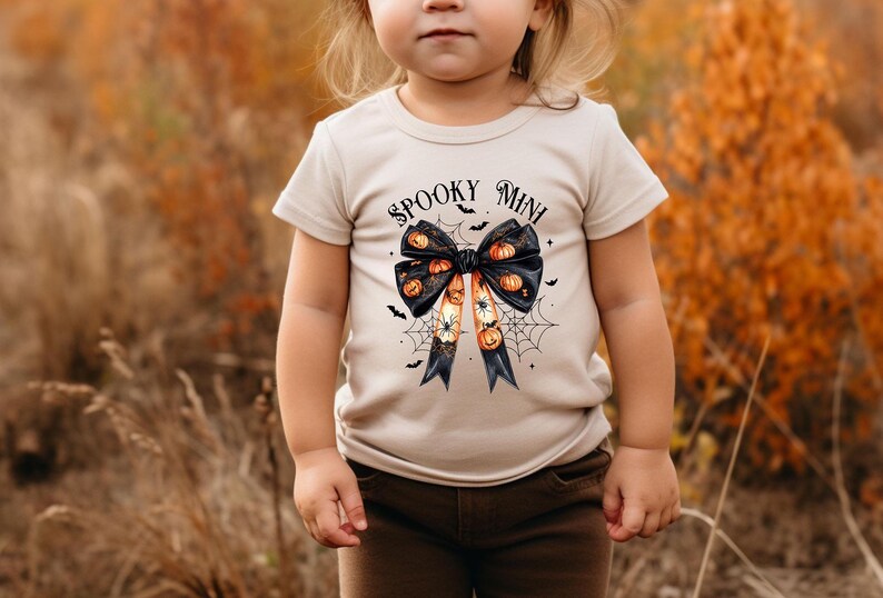 Op de afbeelding: Een peuter draagt een beige T-shirt met een zwarte strik en de tekst "Spooky Mini" erop gedrukt. De strik is versierd met oranje en zwarte pompoenen. De peuter staat in een veld met hoog bruin gras.