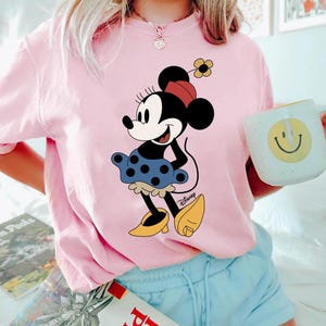 May include: Pink t-shirt featuring a cartoon image of Minnie Mouse wearing a blue polka dot skirt and yellow shoes. The shirt has the word "Disney" printed in the bottom right corner.