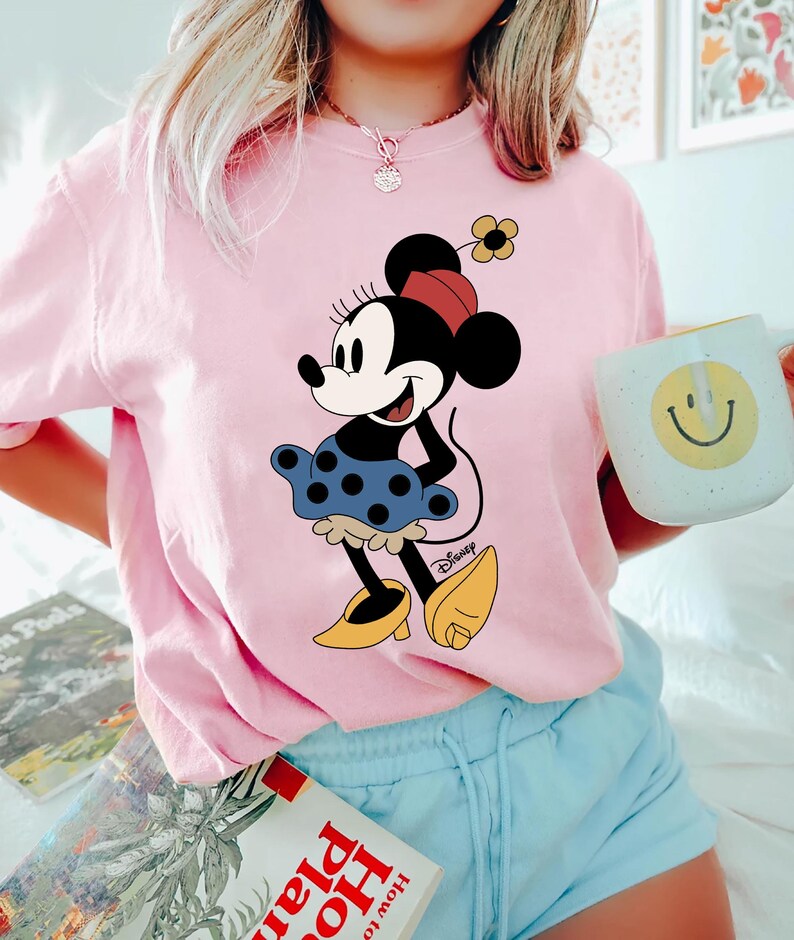 May include: Pink t-shirt featuring a cartoon image of Minnie Mouse wearing a blue polka dot skirt and yellow shoes. The shirt has the word "Disney" printed in the bottom right corner.