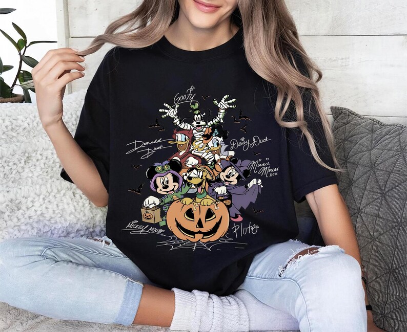Retro-style Halloween design featuring Mickey Mouse, Minnie, Goofy, Donald, Daisy, and Pluto in costumes around a large jack-o'-lantern with bats and spiderwebs. Perfect for Disney Halloween party trips and fall-themed outfits.