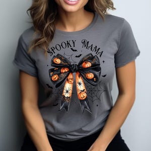 Op de afbeelding: Grijs T-shirt met korte mouwen met een zwarte strik met oranje pompoenen en spinnen erop. De tekst "Spooky Mama" is op het shirt gedrukt.